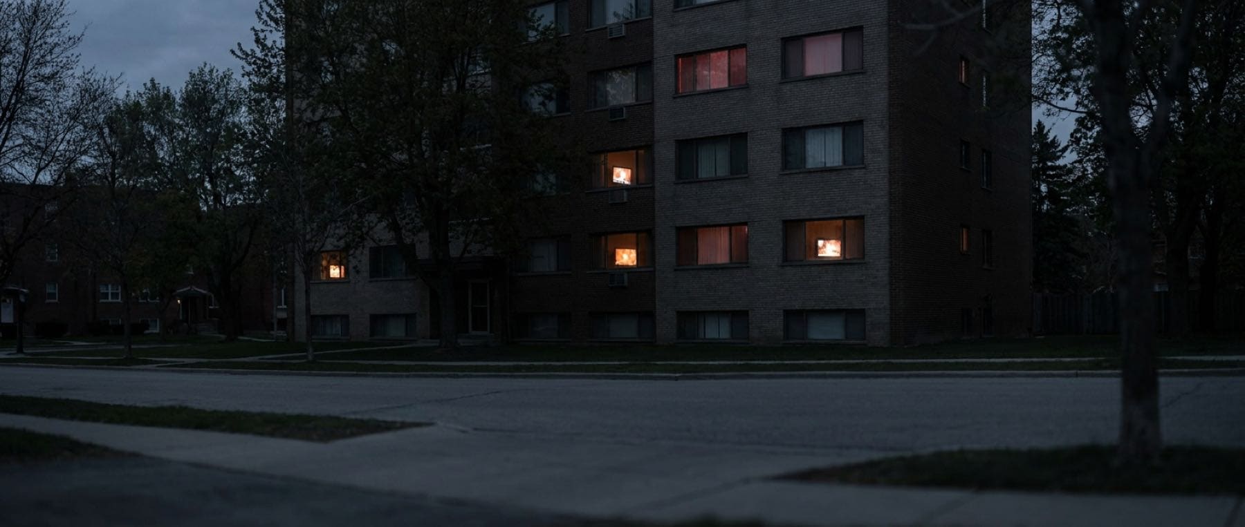 An apartment block at dusk, television light in several windows